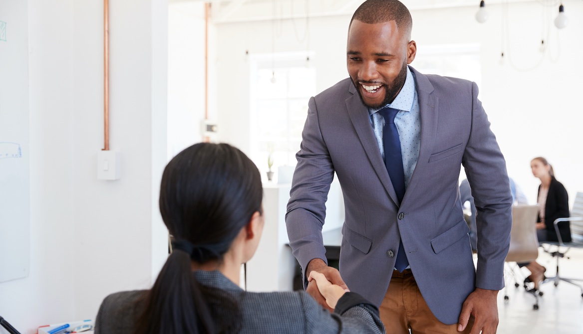 HR manager greeting and shaking hands with seated female job candidate at interview