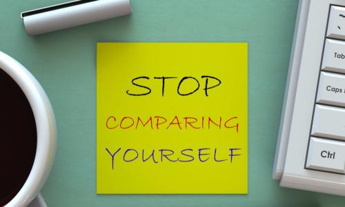 yellow sticky note that says stop comparing yourself on a green desk next to coffee cup and keyboard