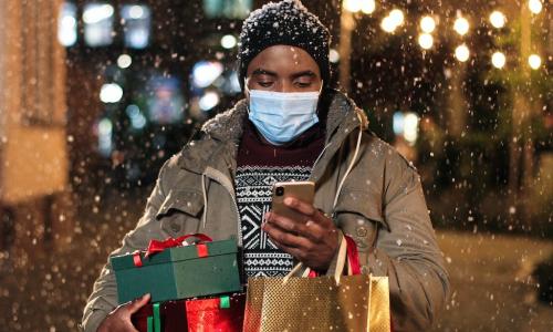 African American man wearing mask looks at smartphone while carrying holiday shopping in snow