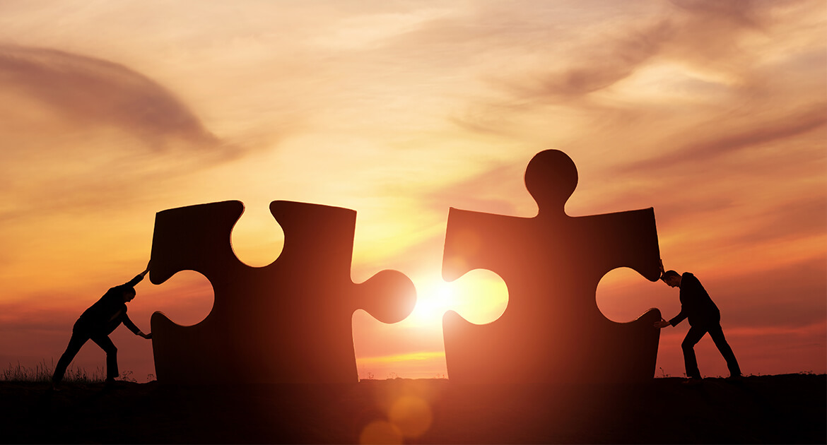 two business men push together two jigsaw puzzle pieces against a backdrop of a sunset