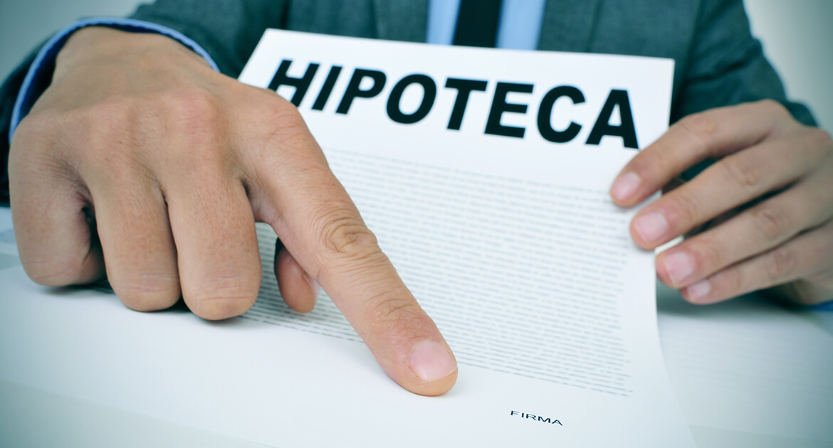 young caucasian man wearing a grey suit sitting at his office desk showing a document with the word hipoteca mortgage loan contract in spanish and where the signer must sign