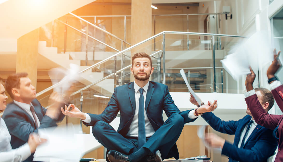 young businessman sits in meditation pose on table while colleagues shout and wave papers at him