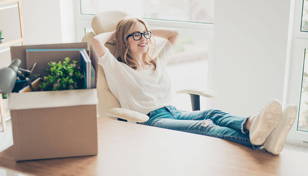 young businesswoman with box of belonging sits with feet up on new desk