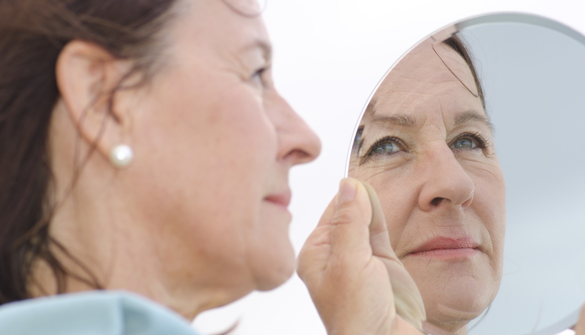 mature woman in pearl earrings looks thoughtfully at her reflection in a hand mirror