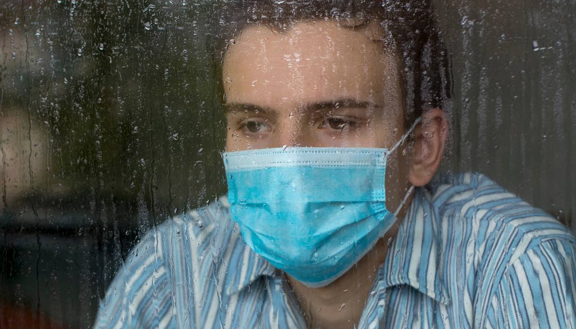 sad man wearing surgical mask looking out window in rain