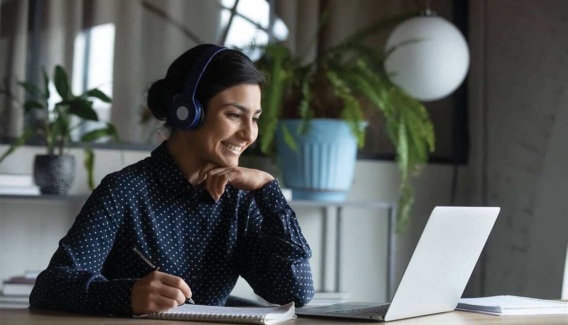 Young businesswoman takes notes while listening to headphones and watching laptop screen