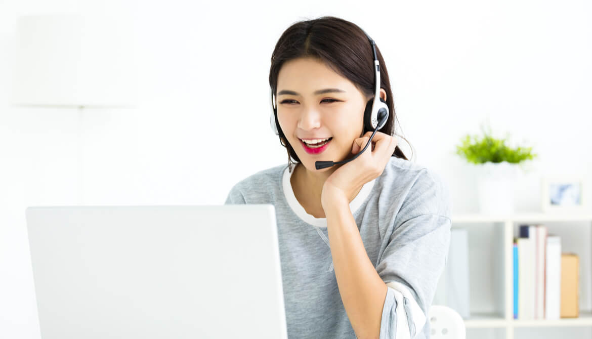 smiling service representative of Asian descent talks on headset while partipating in online training on laptop