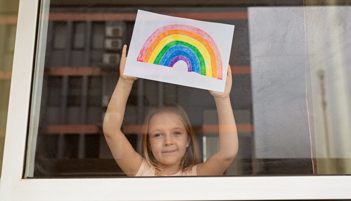 child in window holding up colored drawing of rainbow of hope