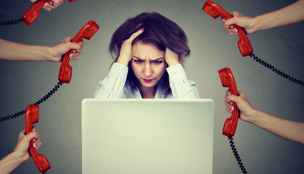 stressed businesswoman holding head while working on laptop surrounded by hands holding out many red telephone receivers