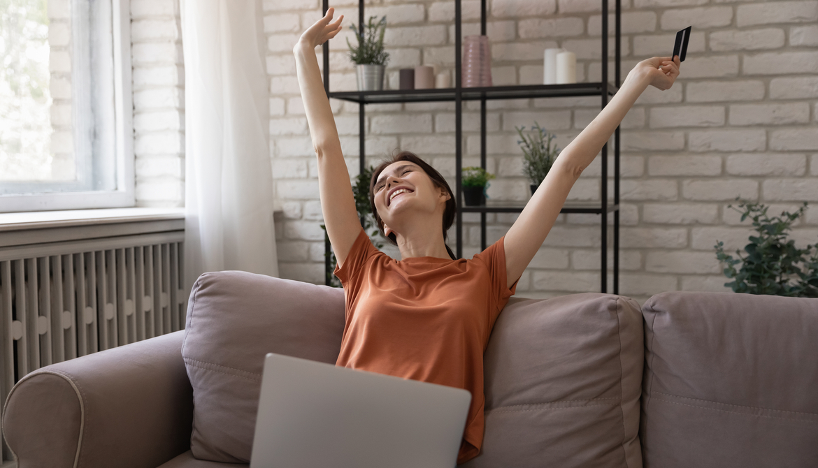 woman on couch with arms up in celebration and laptop and credit card