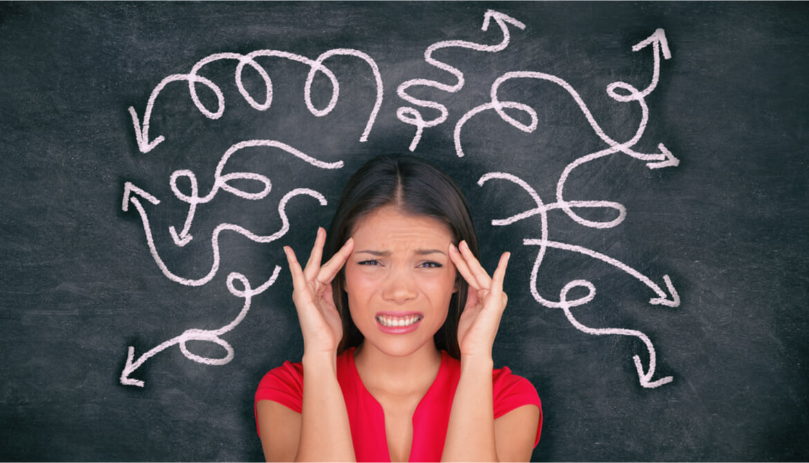 confused and stressed young businesswoman holding her head with tangled arrows drawn on chalkboard behind her
