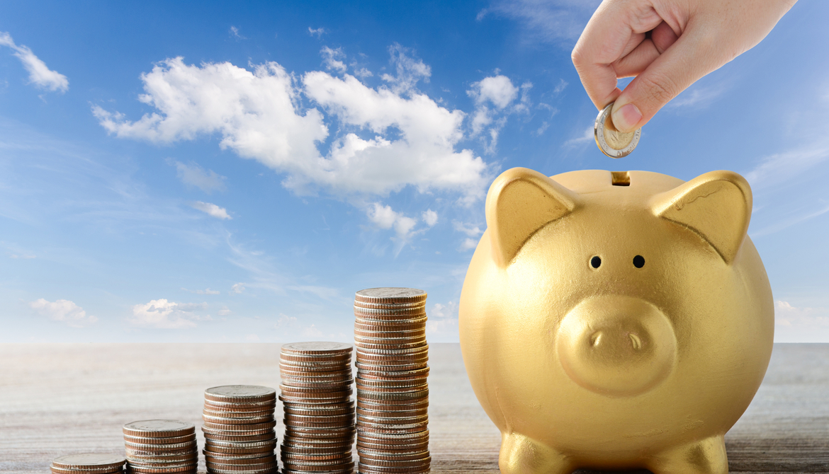 hand depositing coin in piggy bank next to stacks of coins on blue sky