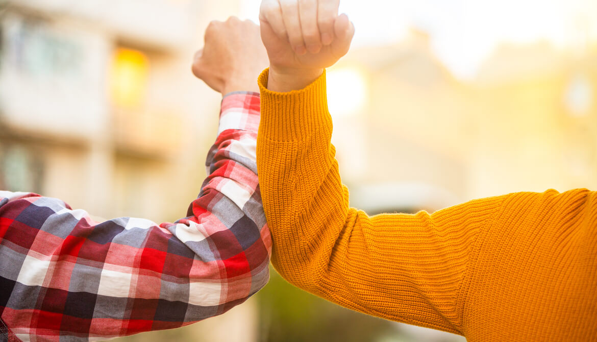 person in plaid shirt elbow bumps person in gold sweater in pandemic greeting