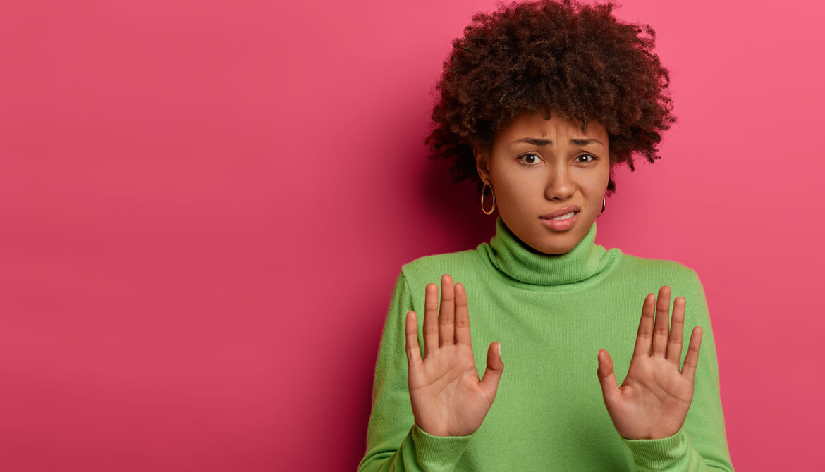 black woman in a green sweater on a pink wall putting her hands up, rejecting something