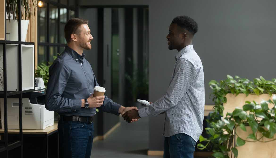 White and black male coworkers shake hands and smile at each other holding cups of coffee in an office