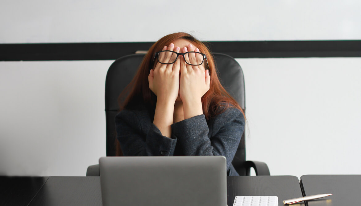 stressed business woman covering her eyes while sitting at desk feeling burnout