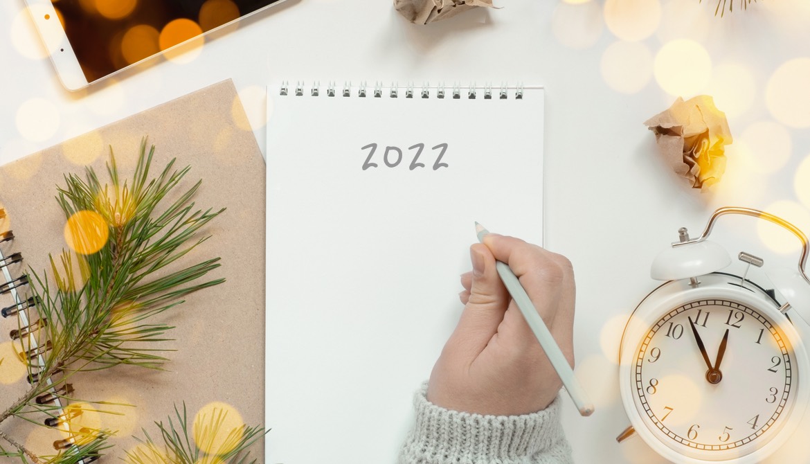 woman’s hand writing 2022 on notebook laying on desk surrounded by alarm clock phone and crumpled papers