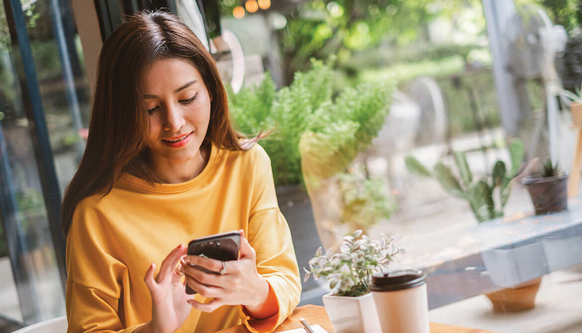 young woman in yellow shirt using smartphone in a café with lots of plants