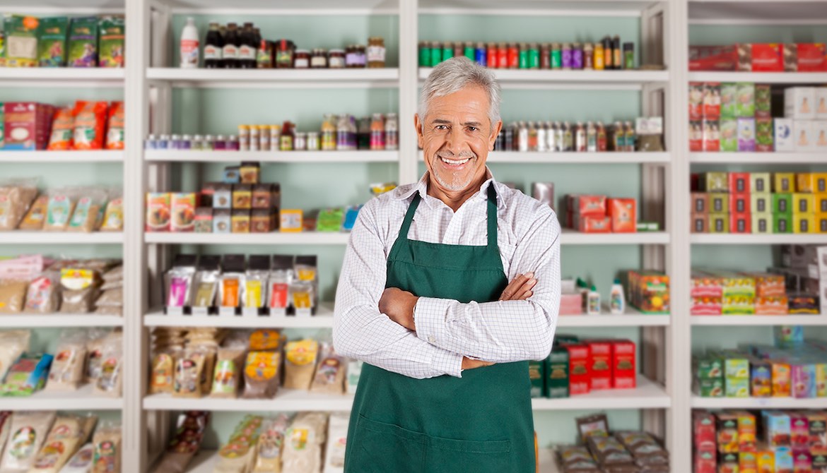 smiling silver haired shop owner in green apron stands in front of store shelves stocked with colorful items