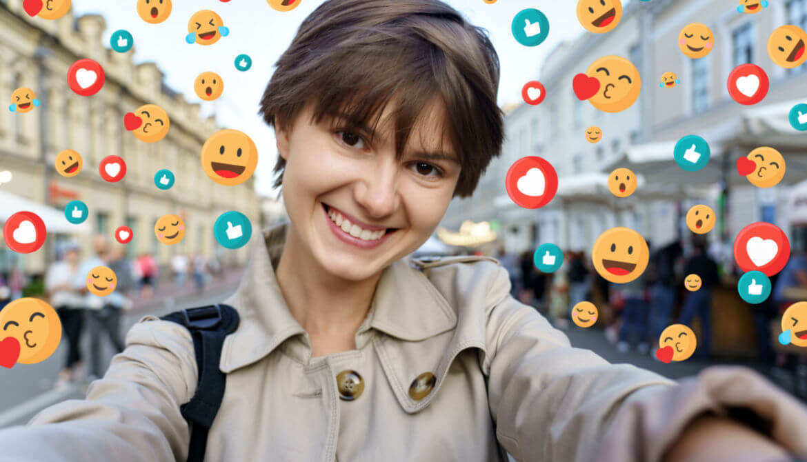 smiling young blogger taking social media selfie in front of city street surrounded by many likes and reaction emojis