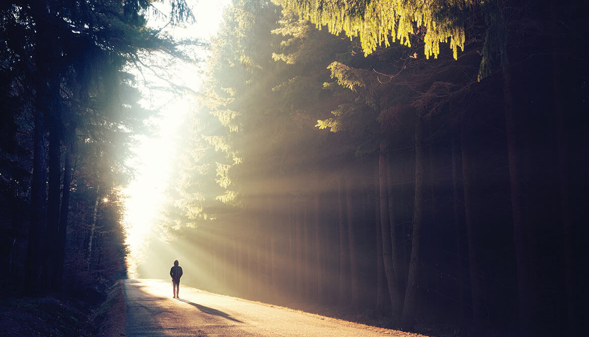 person walking along forest path with sun shining brightly in the distance 