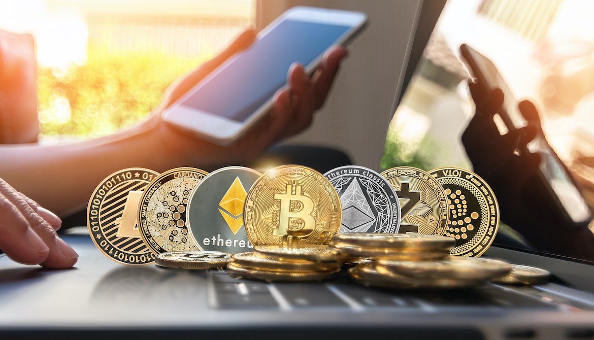 hand holding smartphone next to laptop keyboard covered in cryptocurrency coins