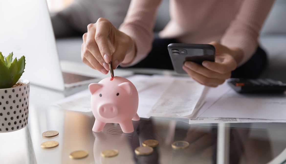 woman dropping a coin into a piggy bank while working on budget at her desk