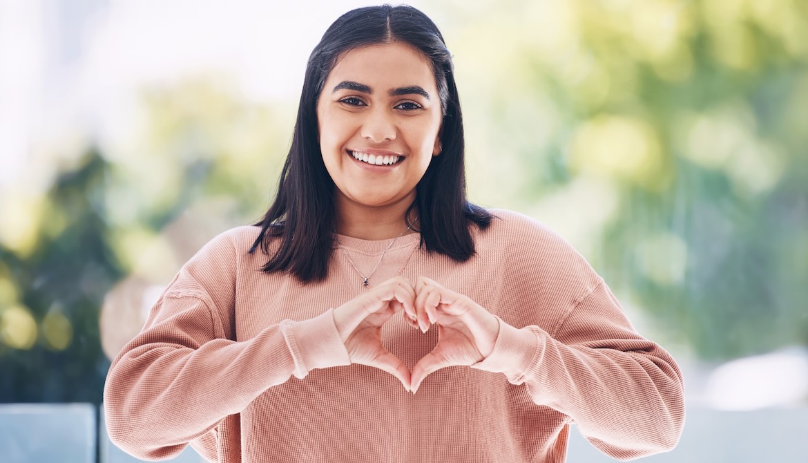 young Gen Z woman in pink sweatshirt making a heart shape with her hands