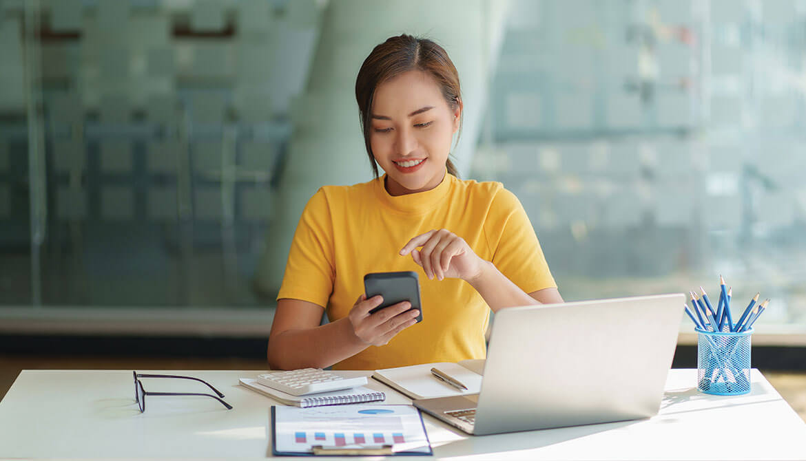 young woman uses online banking on her phone while reviewing financial reports