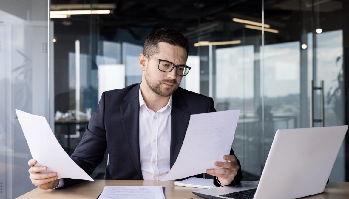 a businessman at a desk reviews with a perplexed expression on his face reviews his executive benefits plan on various papers