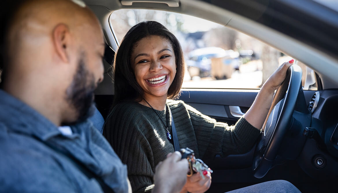 smiling woman sitting in driver seat handing the keys to male passenger in a car
