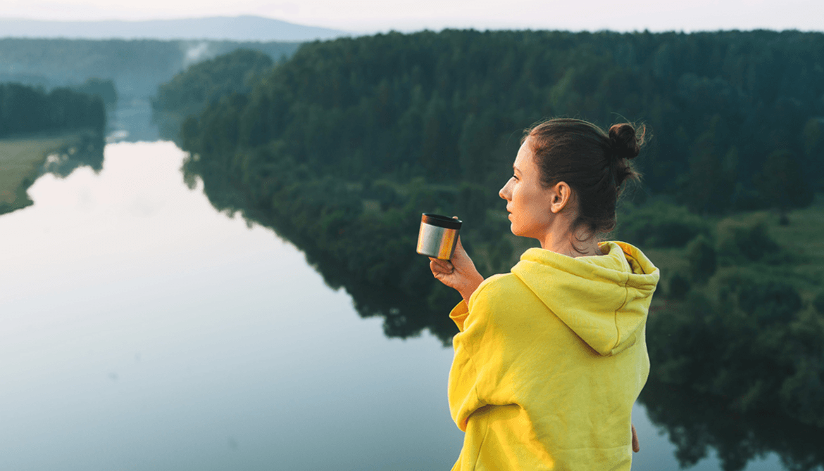 woman in yellow hoodie looking out over a river in a valley while drinking from her cup