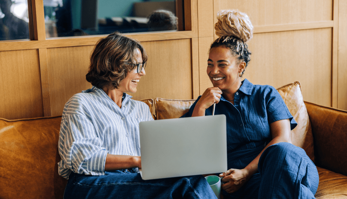 two women smiling sitting on a couch looking at a laptop