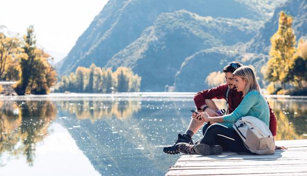 happy couple using smartphone on wooden dock above lake in front of mountains