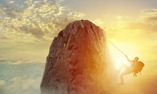 A mountain climber about to reach the top of a mountain peak in front of a cloudy sunset sky 