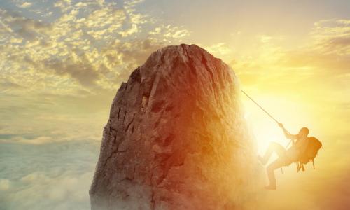 A mountain climber about to reach the top of a mountain peak in front of a cloudy sunset sky 