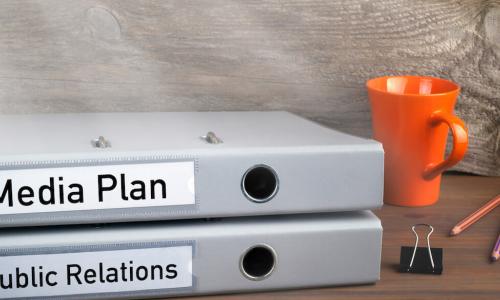 Folders on a desk labeled “media plan” and “public relations”, stacked next to pencils and a coffee mug