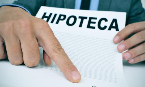young caucasian man wearing a grey suit sitting at his office desk showing a document with the word hipoteca mortgage loan contract in spanish and where the signer must sign
