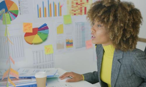 Woman sitting at working desktop with charts on wall and doing her job on computer looking serious