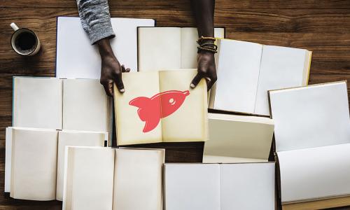 many open blank books on a table with a person holding open a book with a red rocket illustration inside