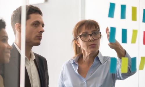 three diverse people discuss information written on colorful sticky notes on a glass wall