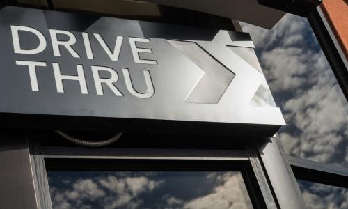 metal drive thru sign on a windowed exterior wall displaying reflection of clouds in the sky