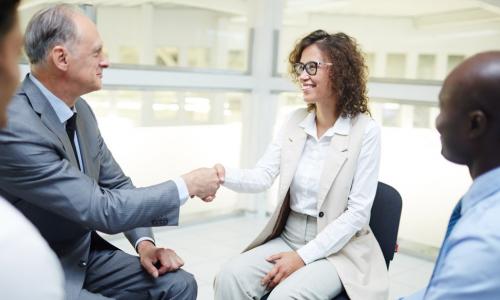 young female director shakes hands with an older executive while a diverse group looks on