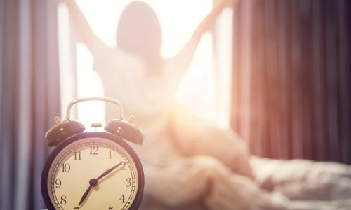 alarm in front of woman sitting up in bed and stretching in front of sunny window