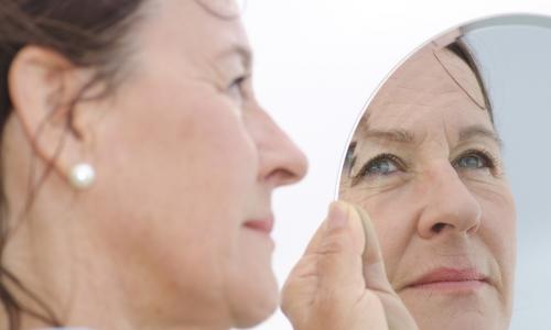 mature woman in pearl earrings looks thoughtfully at her reflection in a hand mirror