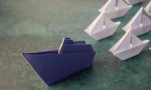 blue origami paper boat leads fleet of smaller white paper boats