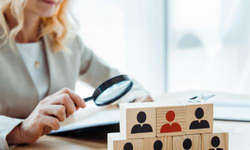 Female HR professional examines pyramid of wooden blocks representing job candidates with a magnifying glass