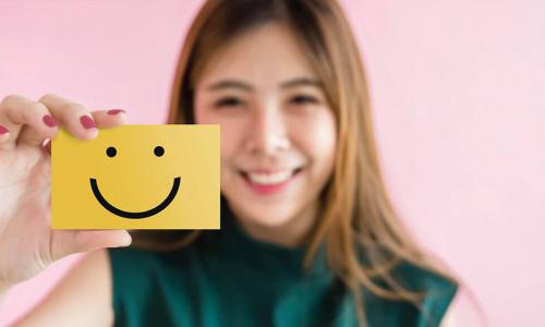 smiling young Asian woman in green shirt holds up yellow notecard with a smiley face drawn on it