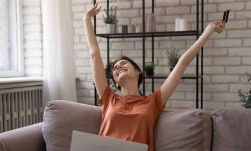 woman on couch with arms up in celebration and laptop and credit card