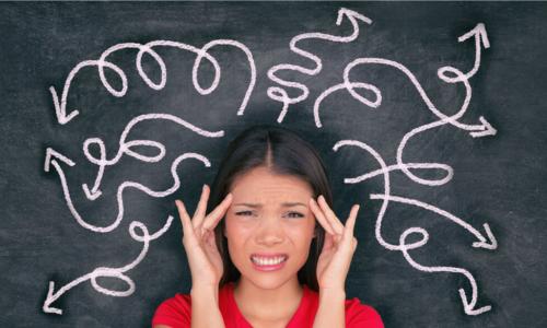 confused and stressed young businesswoman holding her head with tangled arrows drawn on chalkboard behind her